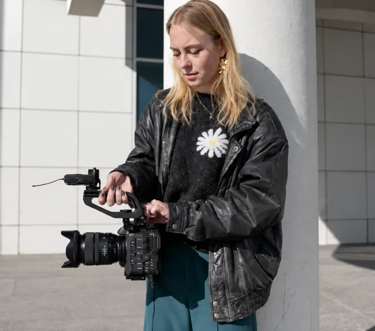 A young filmmaker with a cinema camera leaning against a pillar in the sunshine